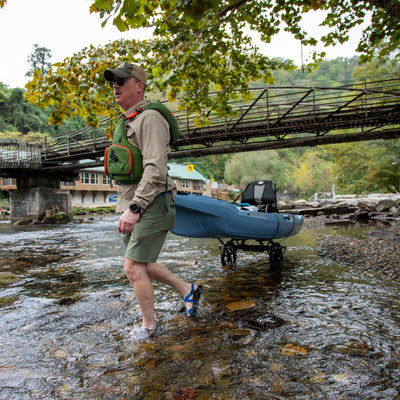 Man walking with a kayak through a shallow stream near a bridge